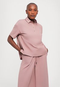 Lys rosa polo skjorte med korte ermer, krage og tre knapper; kombinert med matchende høyt liv bukser med sidelommer.