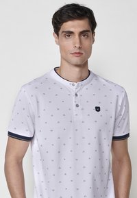 Balta polo marškinių su mygtukais Henley iškirpte, dailiu mažų trikampių geometriniu raštu. Tamsiai mėlynos akcentai ant rankovių ir apykaklės.
