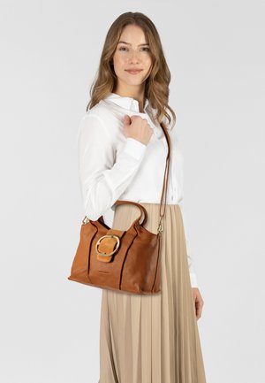 Bruine leren handtas met een afgeronde gouden hardwareaccent, zachte textuur en dubbele banden. Gecombineerd met een model in een witte blouse en een geplooide rok.