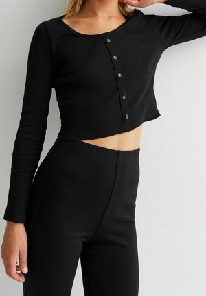 Femme portant un crop top noir à manches longues avec des boutons sur le devant et un pantalon noir taille haute sur un fond uni.