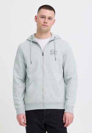 Jonge man met kort haar, gekleed in een lichtgrijze hoodie met rits en tekst op de borst, en zwarte broek, staand tegen een effen witte achtergrond.