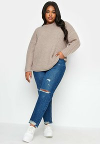Maglione a maglia marrone chiaro con una vestibilità ampia, motivo testurizzato e colletto rotondo, abbinato a jeans blu strappati e scarpe da ginnastica bianche.