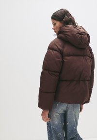 Braune Pufferjacke mit Kapuze, gequiltetem Design und elastischen Bündchen. Das Modell trägt ausgewaschene blaue Jeans.