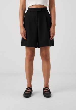 Person iført sorte skræddersyede shorts til knæet, hvid cropped top og sorte Mary Jane-sko stående på en ensartet lys baggrund.
