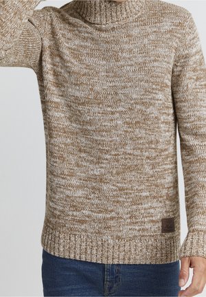 Pull à col roulé tricoté chiné beige et blanc porté avec un jean bleu foncé, montrant un motif texturé et des poignets et un ourlet côtelés.