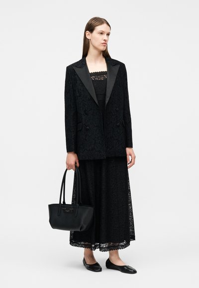 Γυναίκα ντυμένη με μαύρο φόρεμα μέσης από δαντέλα, μαύρο σακάκι με υφή και σατεν πέτα, μαύρα μπαλαρίνες, κρατώντας μια μαύρη τσάντα χειρός με διπλές λαβές.