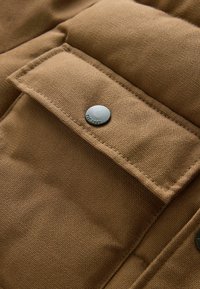 Pruun kangast tasku tekstuuriga pinnaga, mille kinnihoidmiseks on ümmargune metallne nööp, rõhutades õmmeldud servi ja puhast disaini.