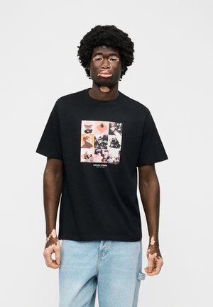 Νέος άνδρας με λεύκη φοράει μαύρο μπλουζάκι με γραφικά και ανοιχτόχρωμο τζιν, στέκεται μπροστά σε απλό ανοιχτό γκρι φόντο.