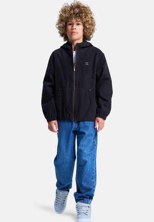 Czarna kurtka z kapturem zapinana na zamek, z elastycznymi mankietami i pasem, w zestawieniu z luźnymi niebieskimi dżinsami i jasnoszarymi sneakersami.