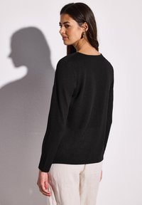 Top nero a maniche lunghe con scollo rotondo. Tessuto morbido con un leggero luccichio. Modella che indossa pantaloni chiari, vista da dietro.