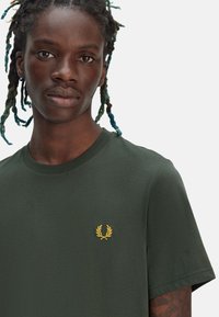 Camiseta de algodón verde oscuro con cuello redondo y mangas cortas, que presenta un logo de laurel bordado en dorado en el lado izquierdo del pecho.