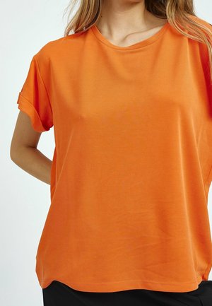 Femme portant un t-shirt à manches courtes orange vif avec des revers, debout avec un bras derrière le dos sur un fond uni.