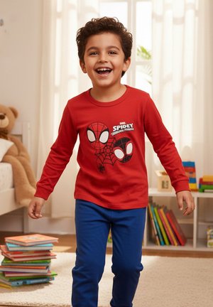 Smiling boy wearing a red Spidey long-sleeve shirt and blue pants stands in a bright room with books and a large teddy bear nearby.