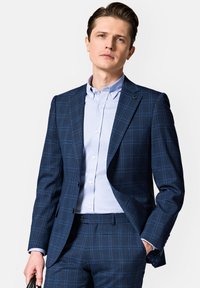 Granatowa marynarka w kratę z dwoma guzikami i szpiczastymi klapami, noszona z jasnoniebieską koszulą z guzikami; gładki materiał, dopasowany krój.