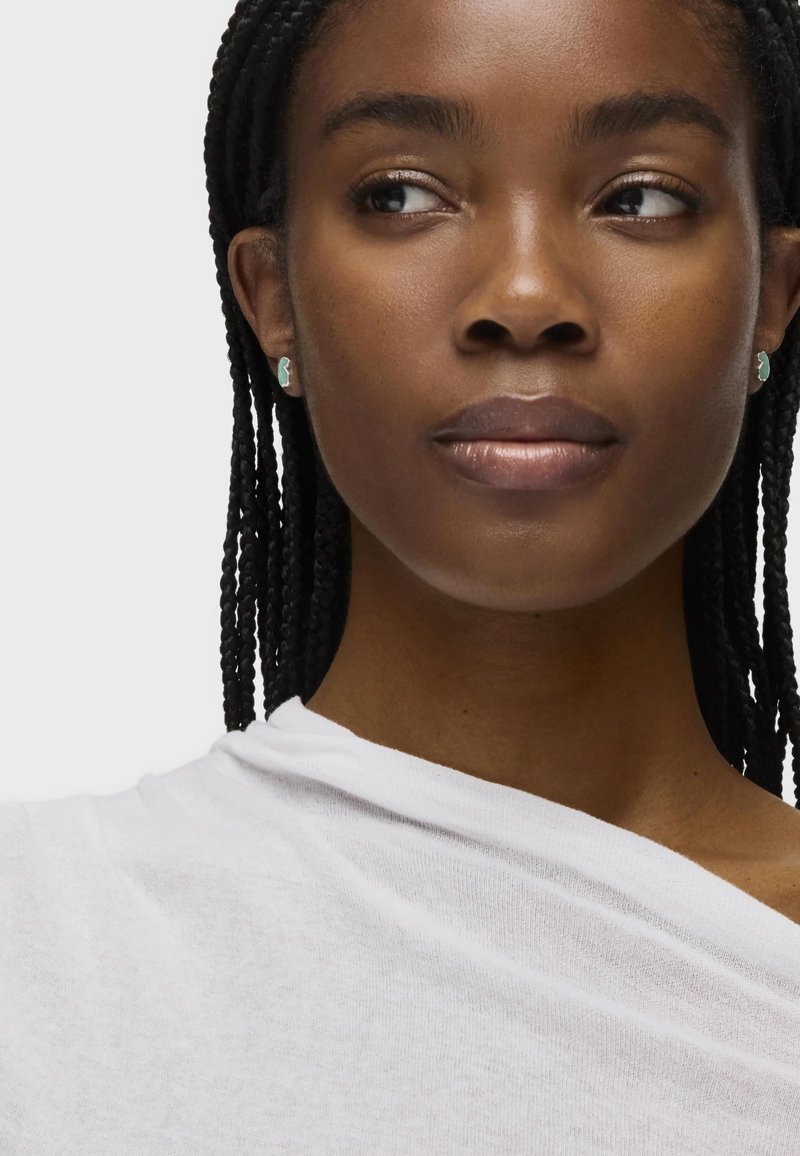 Top blanco, ligero, de tela drapeada con una textura suave. Pendientes delicados con un acento turquesa complementan un look de maquillaje natural.
