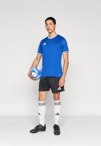 Camiseta deportiva azul con ribetes blancos, pantalones cortos negros, calcetas blancas hasta la rodilla con tres rayas negras y zapatos de fútbol negros, sosteniendo una pelota multicolor.