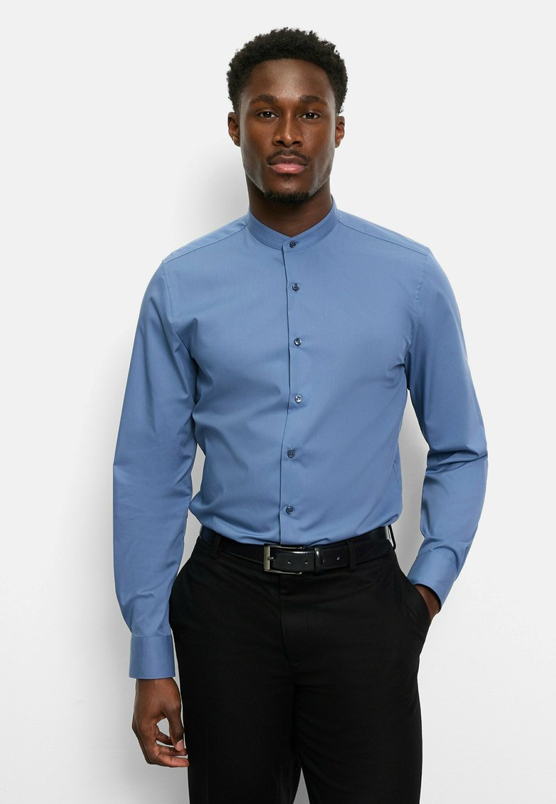 Langärmliges, hellblaues Hemd mit Stehkragen, Knopfverschluss und glatter Textur; kombiniert mit schwarzen Hosen und Gürtel.