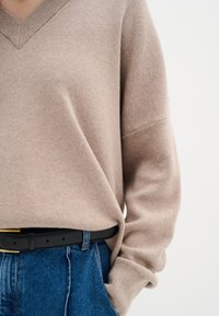 Beżowy sweter w serek z miękkiej dzianiny z luźnymi rękawami, noszony na spodniach dżinsowych z wysokim stanem, zabezpieczony czarnym paskiem.