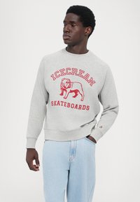 Grå sweatshirt med rød tekst "ICECREAM SKATEBOARDS" og løve-grafik. Har ribbede håndled og søm, båret med lyseblå denimjeans.