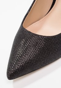 Escarpin noir à bout pointu avec une surface texturée, présentant une finition brillante et une doublure intérieure lisse beige.