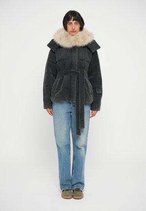 Pufferjack in donkergrijs corduroy met een grote nepbontkraag, een ceintuur in de taille en opgestikte zakken, gecombineerd met lichtblauwe jeans en groene schoenen.