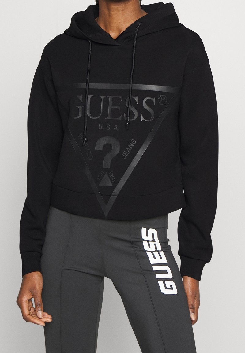 Personne portant un sweat à capuche noir Guess avec un grand logo et des leggings noirs avec un logo vertical blanc Guess sur la cuisse.