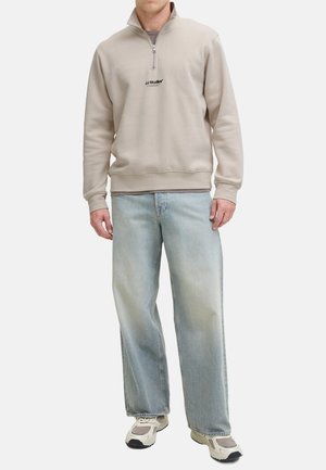 Beige zip-up sweatshirt met een hoge kraag, voorzien van een logo op de borst. Gepaarde met lichtblauwe, wijde jeans en witte sneakers.
