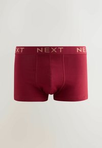 Sarkanas kokvilnas biksītes ar ciešu piegulumu, plaša jostasvieta ar zelta uzrakstu "NEXT". Dizainam ir gluda tekstūra bez rakstiem.