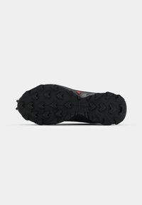 Semelle de chaussure de sport noire en caoutchouc texturé, présentant des motifs de bande de roulement agressifs et un logo de prise centrale en rouge bien en évidence.