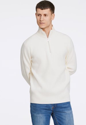 Hvid ribstrikket sweater med høj krave og halv lynlås, parret med blå denim jeans. Stoffet ser ud til at være strikket med en tekstureret overflade.