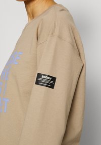 Sudadera beige con cuello redondo, con texto gráfico azul. Incluye una etiqueta de tela negra con información de la marca en la manga izquierda.
