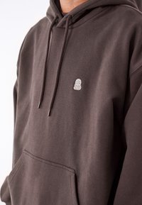 Mörkbrun huvtröja i mjukt tyg, med en ficka fram, dragsnören och en liten broderad logotyp på bröstet.