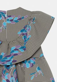 Tkanina w czarno-białą pepitkę z niebieskim i fioletowym haftem kwiatowym. Posiada falbaniasty dekolt i zasuwaną zamek ukrytą.