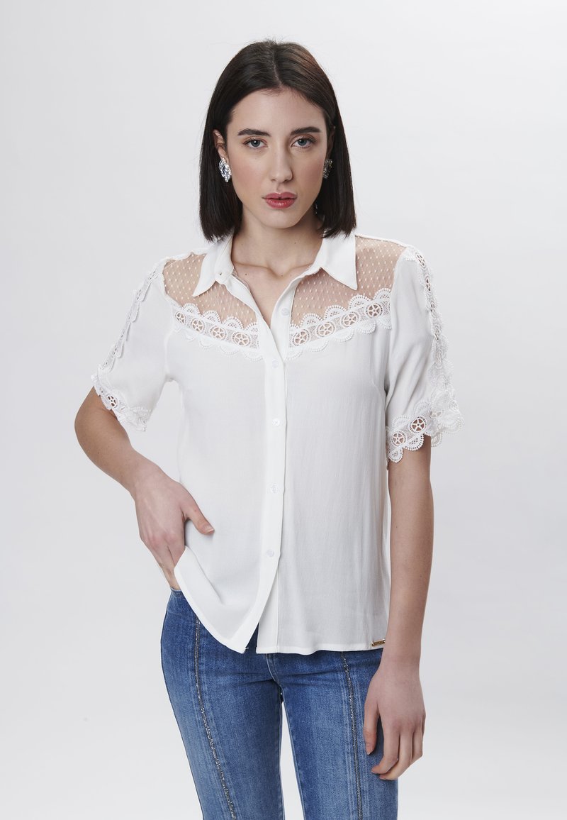 Blusa bianca con pizzo e dettagli in rete sulle spalle e sulle maniche, chiusura frontale con bottoni e maniche corte. Abbinata a jeans in denim blu.