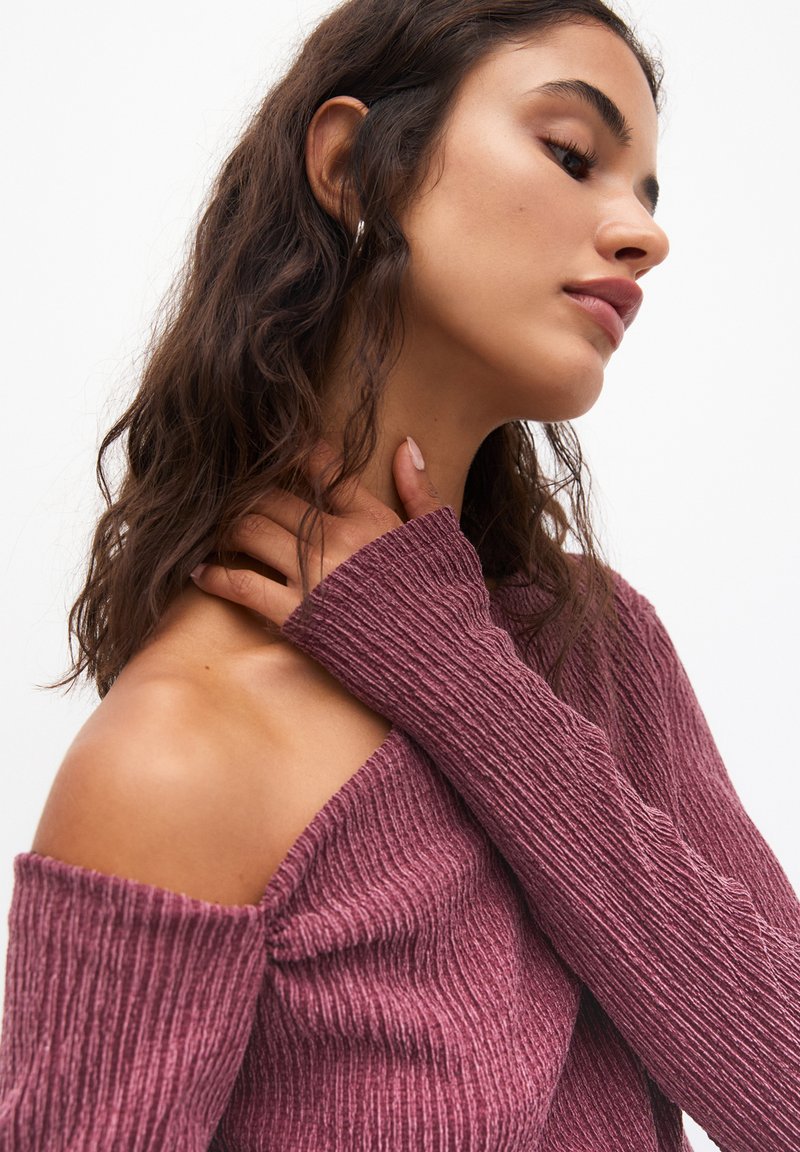 Top a maniche lunghe e spalle scoperte realizzato in tessuto a coste testurizzato di colore bordeaux, con un design aderente e maniche attillate.