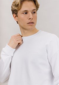 Hvitt sweatshirt i myk stoff med ribbestrikket hals og mansjetter. Har subtil preget tekst på brystet. Modell som forsiktig drar i kragen.