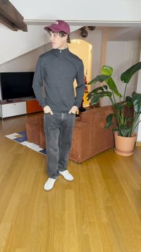 Gray zip-up sweatshirt, black jeans, and white sneakers. Burgundy cap, standing indoors near a potted plant and a brown couch.
