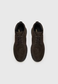 Chaussures à lacets en suede marron avec un bout rond, dessus texturé et semelle en caoutchouc, dotées de lacets gris et d'un logo subtil sur la semelle intérieure.