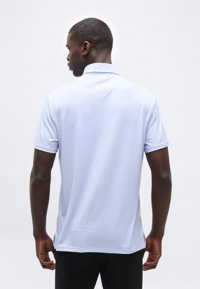 Άνδρας με την πλάτη στραμμένη, φοράει κοντομάνικη πόλο μπλούζα ανοιχτού μπλε χρώματος με υφή και μαύρο παντελόνι, μπροστά σε απλό φόντο.