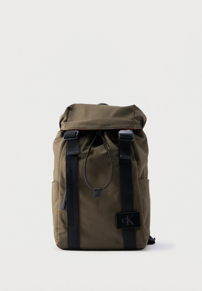 Sac à dos vert olive avec des sangles noires, une fermeture à cordon, et un petit patch carré noir avec le logo "cK" sur la partie inférieure avant.