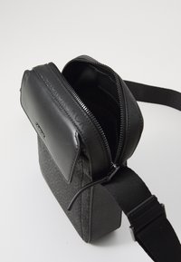 Borsa a tracolla nera in canvas con battente in pelle texturizzata, apertura con zip e tracolla regolabile; presenta slot per carte interne e logo.