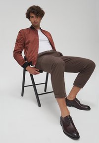 Giubotto bomber marrone con accenti neri, t-shirt bianca, pantaloni marroni arrotolati e scarpe in pelle marrone scuro, seduto su una sedia nera contro uno sfondo neutro.