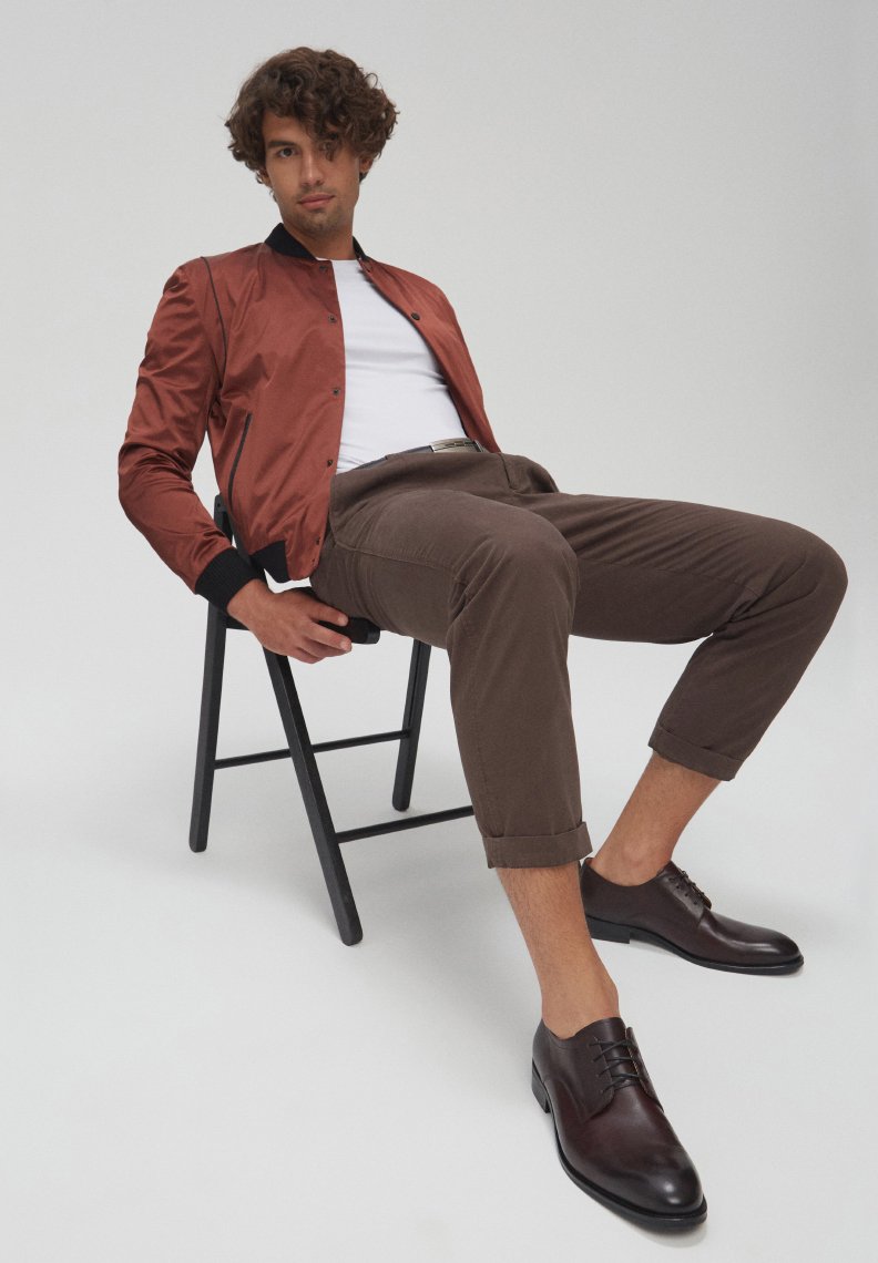 Giubotto bomber marrone con accenti neri, t-shirt bianca, pantaloni marroni arrotolati e scarpe in pelle marrone scuro, seduto su una sedia nera contro uno sfondo neutro.