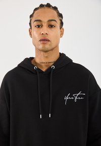 Junger Mann mit geflochtenem Haar trägt einen schwarzen Hoodie mit dem Text "Your Turn" und einem Nackentattoo, das "Believe in yourself" liest, vor einem schlichten Hintergrund.