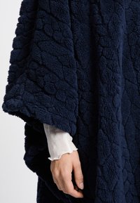 Poncho di pile blu navy con un motivo testurizzato, caratterizzato da un tessuto morbido e peloso e da un design ampio e aperto. Messo in risalto da una camicia bianca sotto.