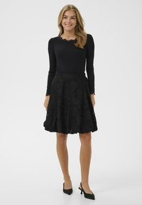 Nasmejana žena z dolgimi blond lasmi, oblečena v črno majico z dolgimi rokavi, teksturirano črno krilo do kolen in črne pete, na belem ozadju.