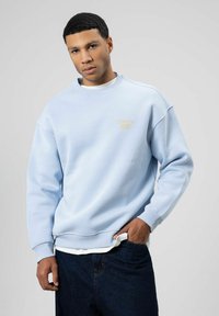 Homme aux cheveux courts noirs portant un sweat-shirt bleu clair et un jean foncé, debout avec une main dans la poche, sur un fond gris clair.