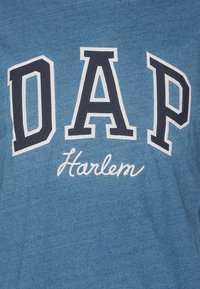 Modra bombažna majica s boldnimi mornarskimi črkami "DAP" in besedo "Harlem" v beli pisanici, z mehkim, teksturiranim videzom tkanine.