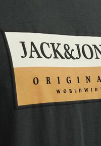 Grøn bomulds-t-shirt med et stort grafik i creme og brunt med "JACK&JONES" og "ORIGINAL" i fed sort tekst. Glat tekstur.