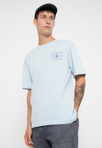 Jonge man in een lichtblauw T-shirt met paarse tekst en afbeelding "ONLY&SONS NOODLES", donkere jeans, een zwart pet en een tatoeage op zijn linkervoorarm.